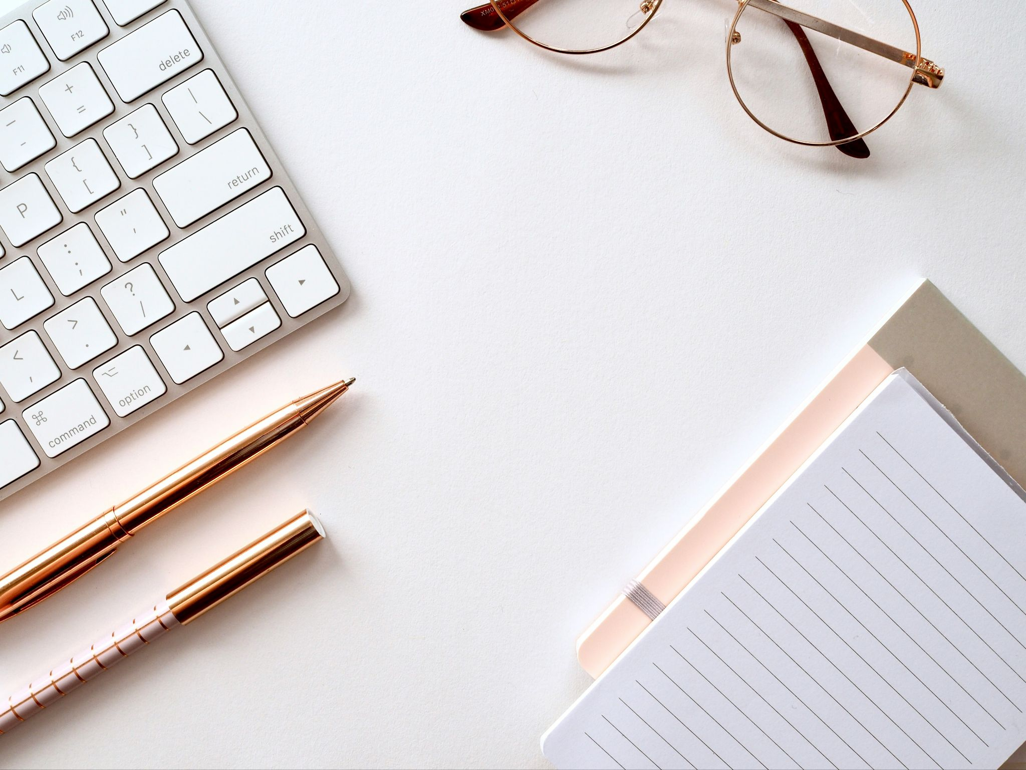 Flat lay of a keyboard, glasses, a pen, and a notepad on a white surface.