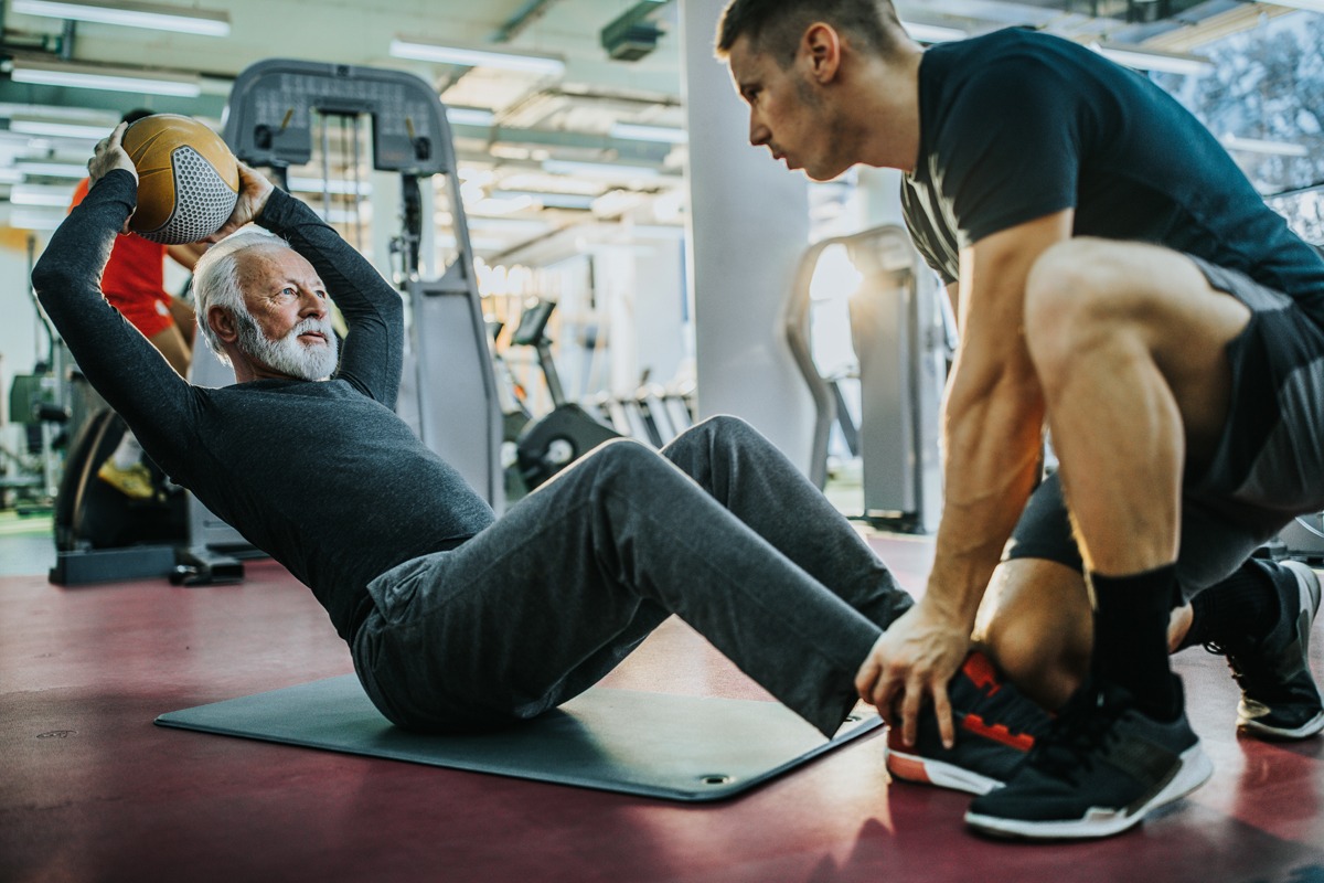 An older man performs a medicine ball exercise while a younger trainer assists him in a gym setting.