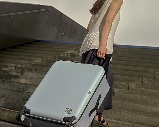 A person in a white top and dark skirt pulls a light blue suitcase up a set of outdoor stairs.