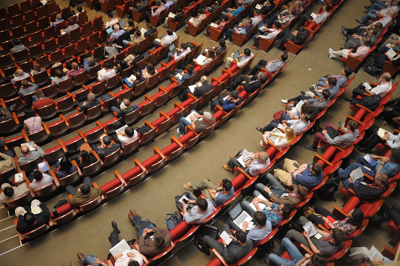Aerial view of a large audience seated in a theater, engaged in a presentation or lecture with laptops and notepads.