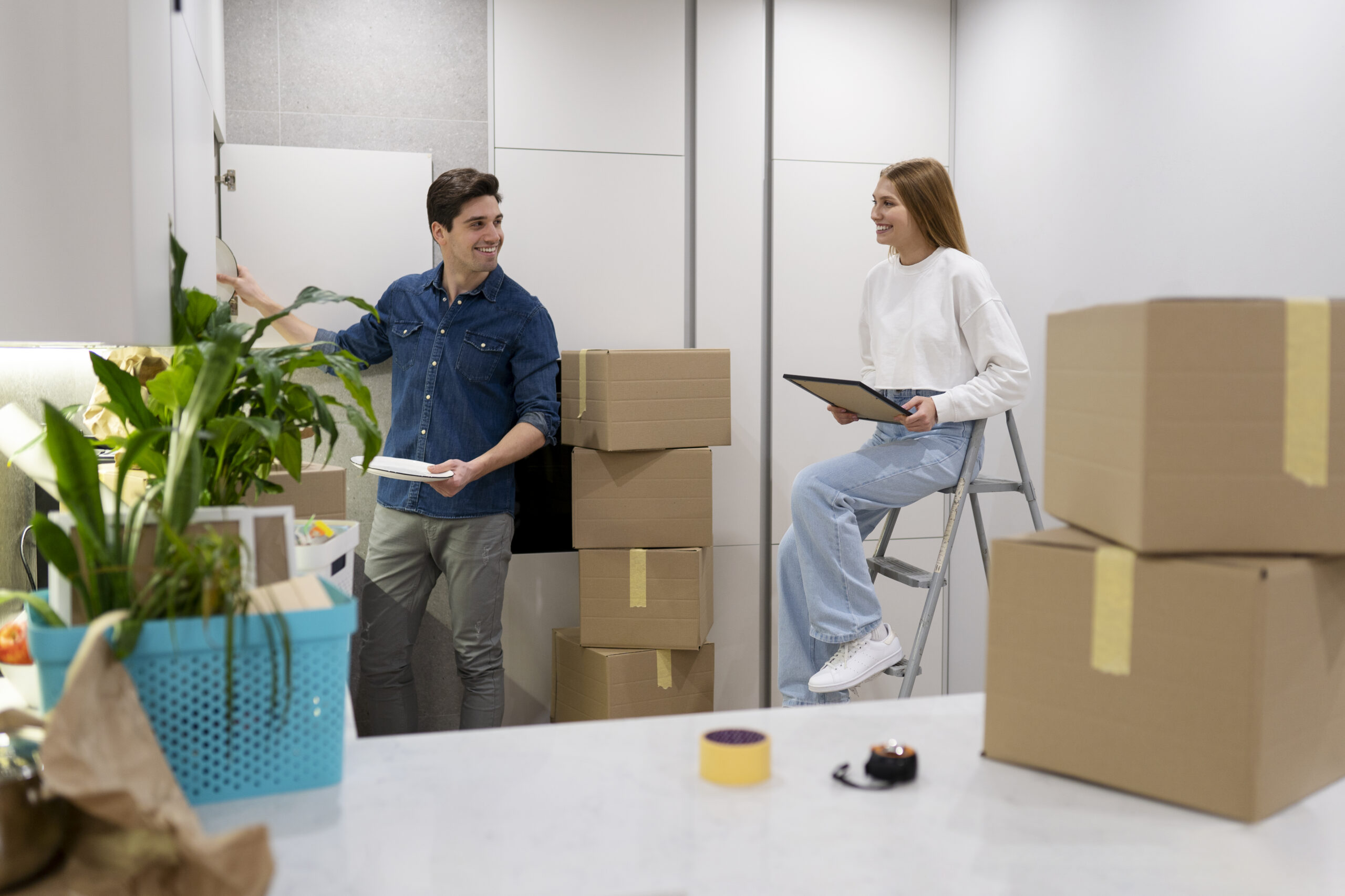 A man and woman organize boxes in a modern kitchen, with plants and a ladder nearby. The woman holds a clipboard.