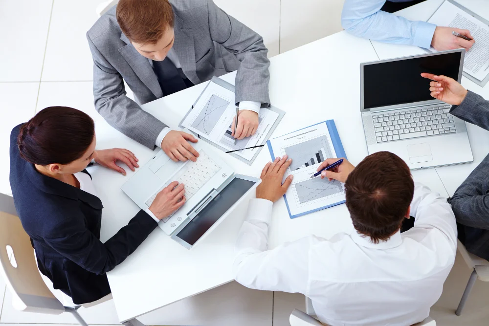 Group of professionals in business attire collaborating at a table with laptops and documents featuring charts and graphs.